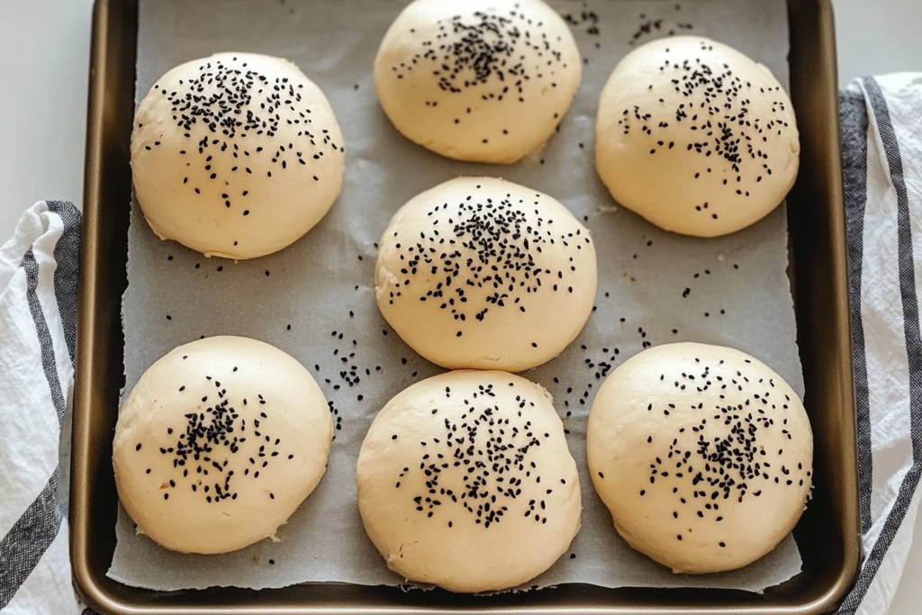 Unbaked dough balls sprinkled with black sesame seeds, evenly arranged on a parchment-lined baking tray with a striped kitchen towel beside it