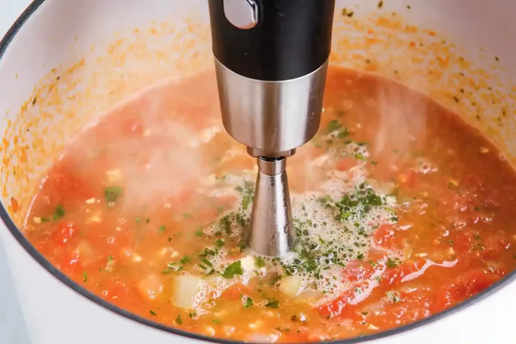 An immersion blender blending the tomato soup with herbs in a white pot, creating a smooth and creamy texture.