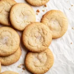 A pile of golden sourdough discard sugar cookies on crinkled parchment paper, lightly dusted with sugar.