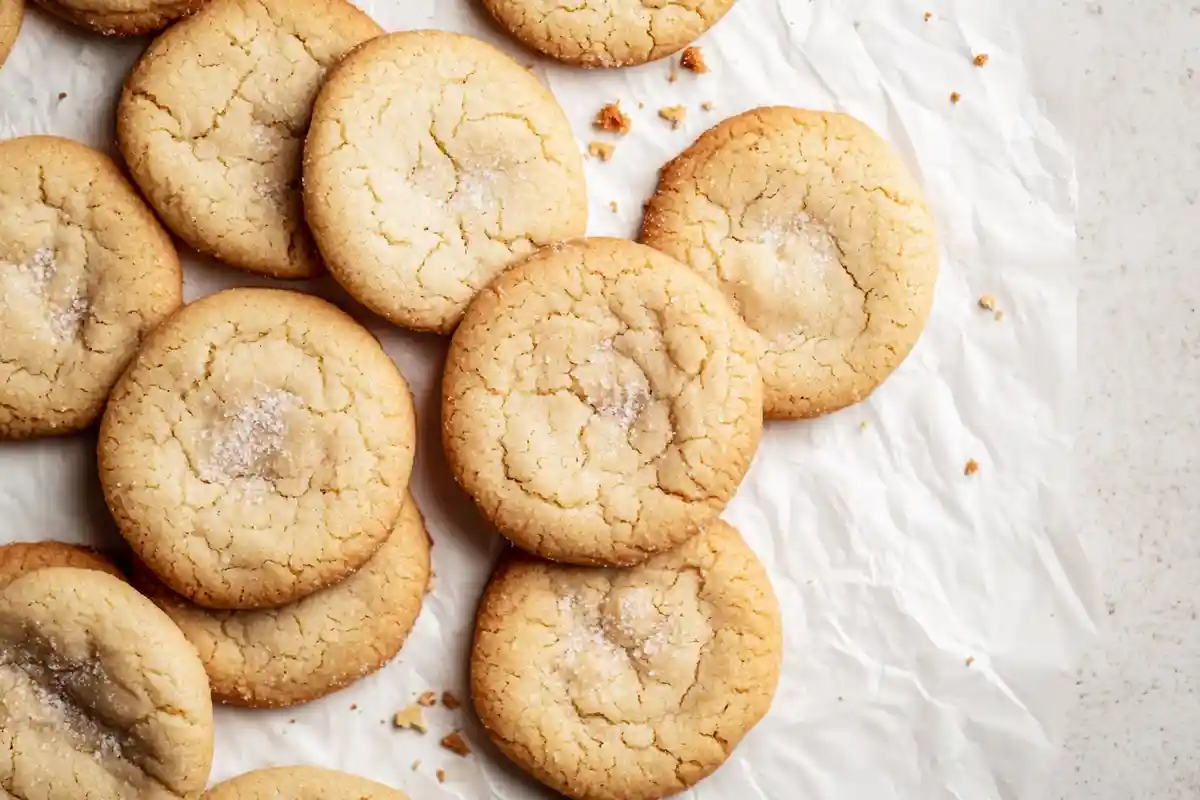 A pile of golden sourdough discard sugar cookies on crinkled parchment paper, lightly dusted with sugar.