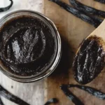 A glass jar filled with glossy homemade vanilla paste, with a wooden spoon coated in the paste, surrounded by fresh vanilla beans on a wooden surface.