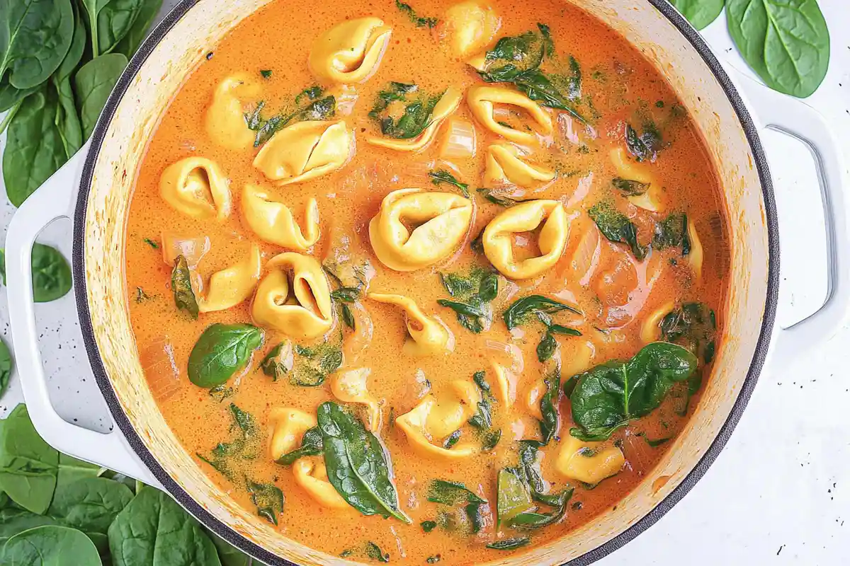 An overhead view of a pot filled with creamy tomato soup, spinach, and cheese tortellini, showcasing vibrant colors and a hearty texture