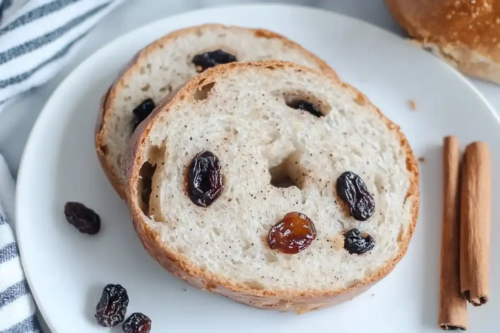 Two slices of sourdough cinnamon raisin bagel on a white plate, surrounded by raisins and cinnamon sticks.