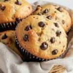 Freshly baked sourdough discard chocolate chip muffins in a linen-lined basket with scattered chocolate chips around.