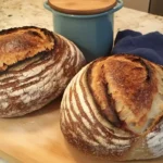 Two artisan sourdough bread loaves with golden crusts and decorative scoring, placed next to a blue ceramic jar and cloth on a kitchen counter.