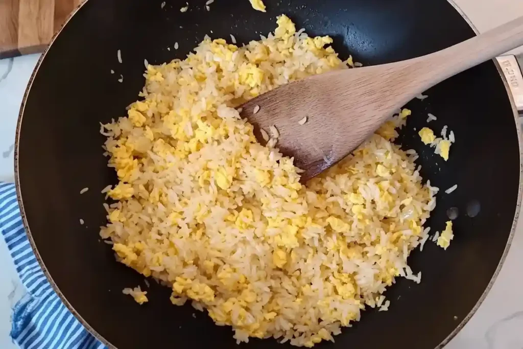 A wooden spoon mixing golden yellow scrambled eggs and fluffy white rice in a black wok.