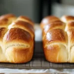 Two beautifully golden-brown sourdough brioche loaves cooling on a wire rack, with a rich, shiny crust and soft, fluffy texture.